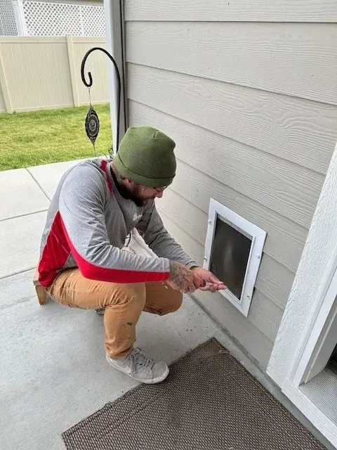 A Mr.Handyman service professional is kneeling down installing a pet door from outside the house.