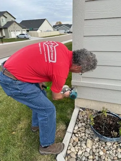 A Mr. Handyman service professional crouches down and uses an electric drill to install siding on a house.