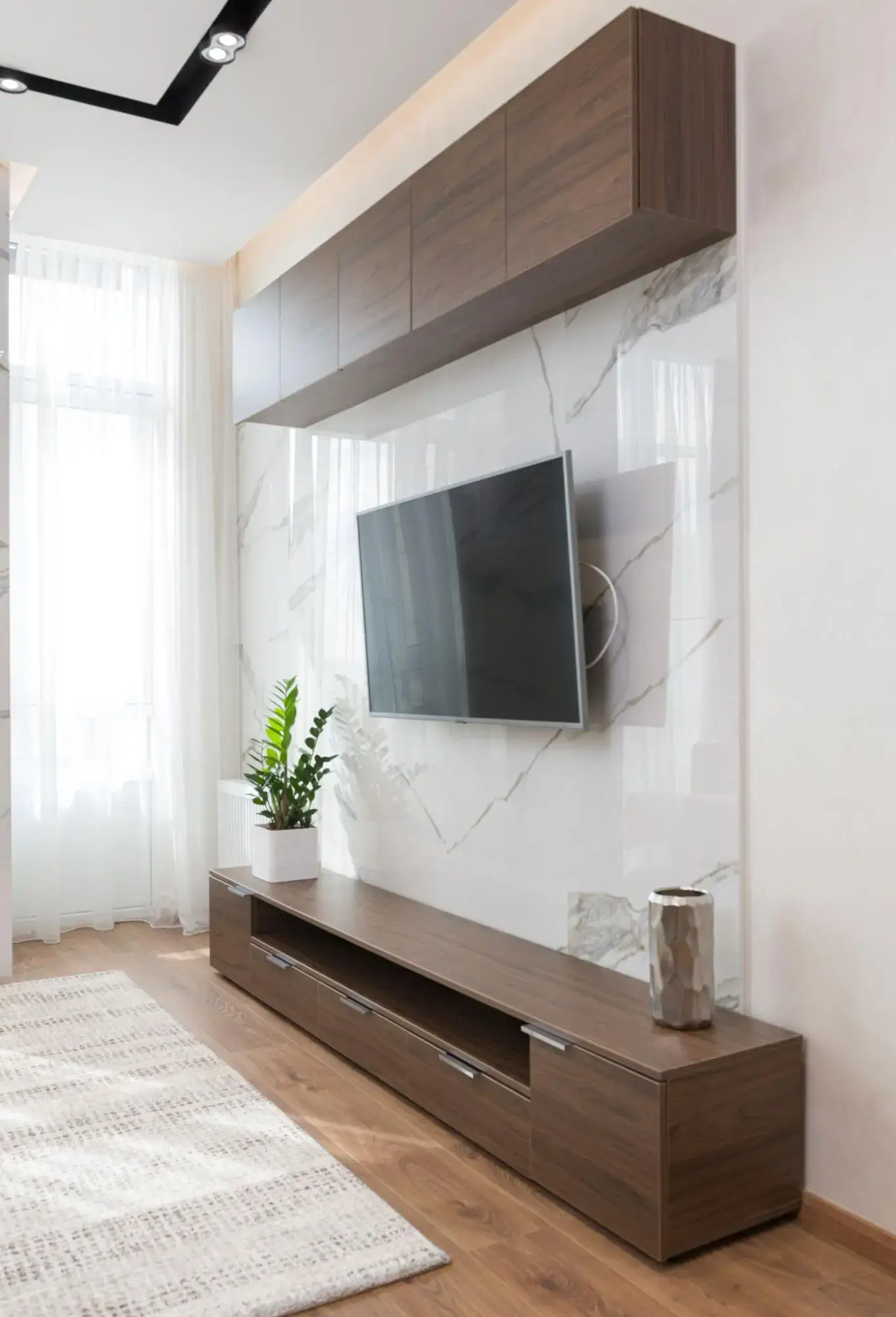 A large tv screen is installed on the wall in a sleek, modern living room. There is a wooden entertainment unit below the tv with a houseplant and vase on it.
