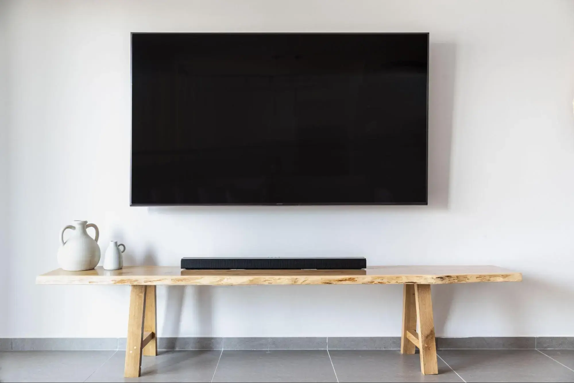 A large flat screen tv is installed on a white wall. A wooden bench with a sound bar and a few vases sits below it.