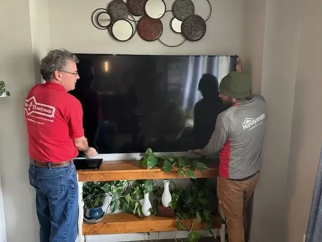 Two Mr. Handyman service professionals install a large flat screen tv over a set of shelves that are holding houseplants.
