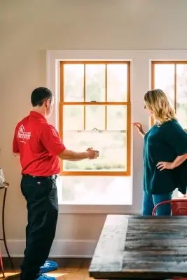 A Mr.Handyman service professional in a red shirt and a blonde woman in a green shirt inspect window frames together.