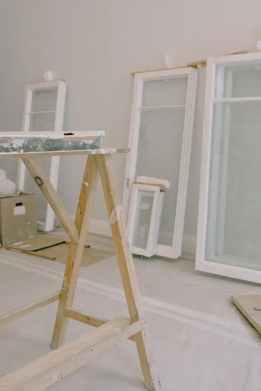 A room under construction with many white windows stacked against the wall. There is a workbench in the foreground.