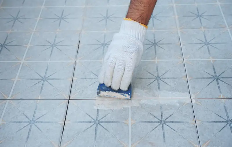 Gloved hand spreading grout between gray patterned tiles.