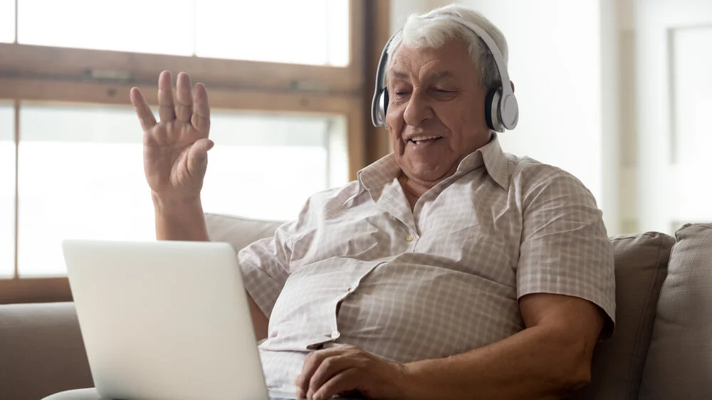 Senior citizen wearing headphones and waving at laptop screen.