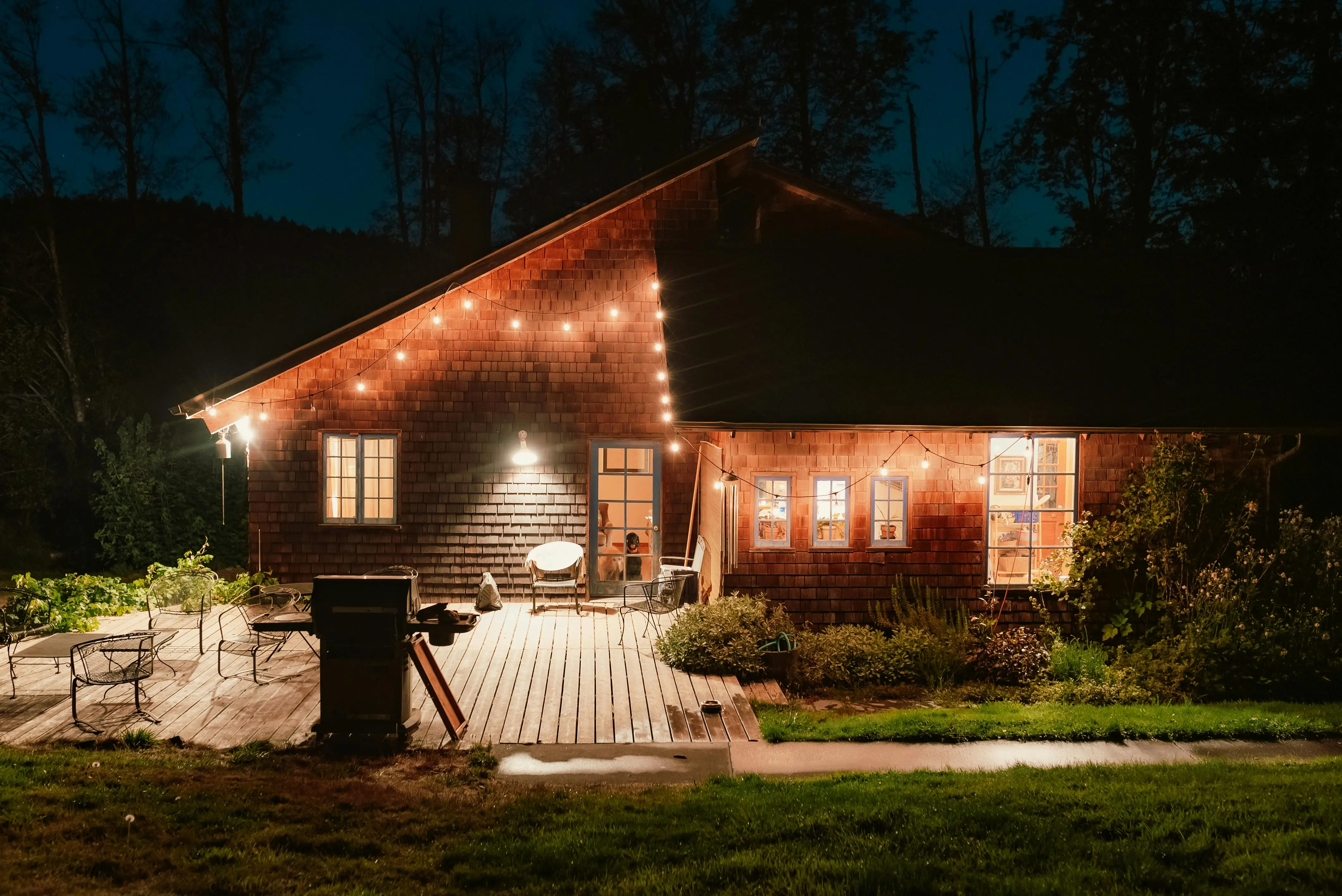 An image of a house with wooden siding surrounded by bushes, trees, and grass. It is lit brightly by outdoor string lights and lights from inside the house. On the outside, chairs, and grill sit on a wooden deck.
