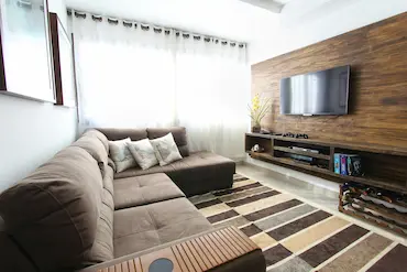 A brown sectional couch sits in front of a wooden-panelled wall. A TV is mounted on the wall, with a small shelf underneath filled with books and video game controllers.