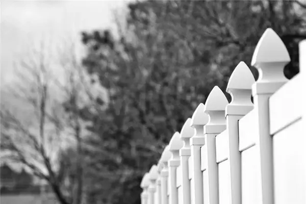 A black and white photo of a vinyl fence with trees in the background.