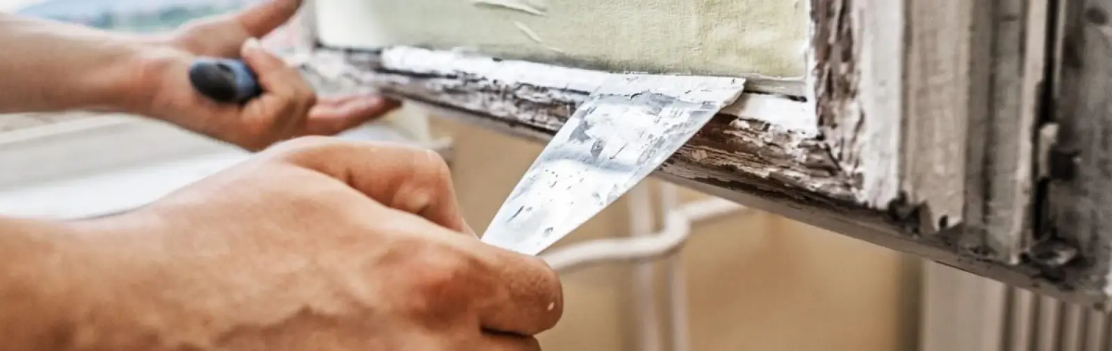 Using a putty knife to scrape deteriorated caulk and paint from an aging wooden window frame.