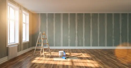 Empty sunlit room with ladder and drywall repair materials.