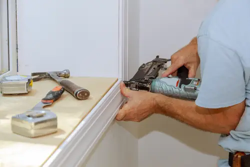 Worker installing window trim with nail gun, hammer, putty knife and tape measures
