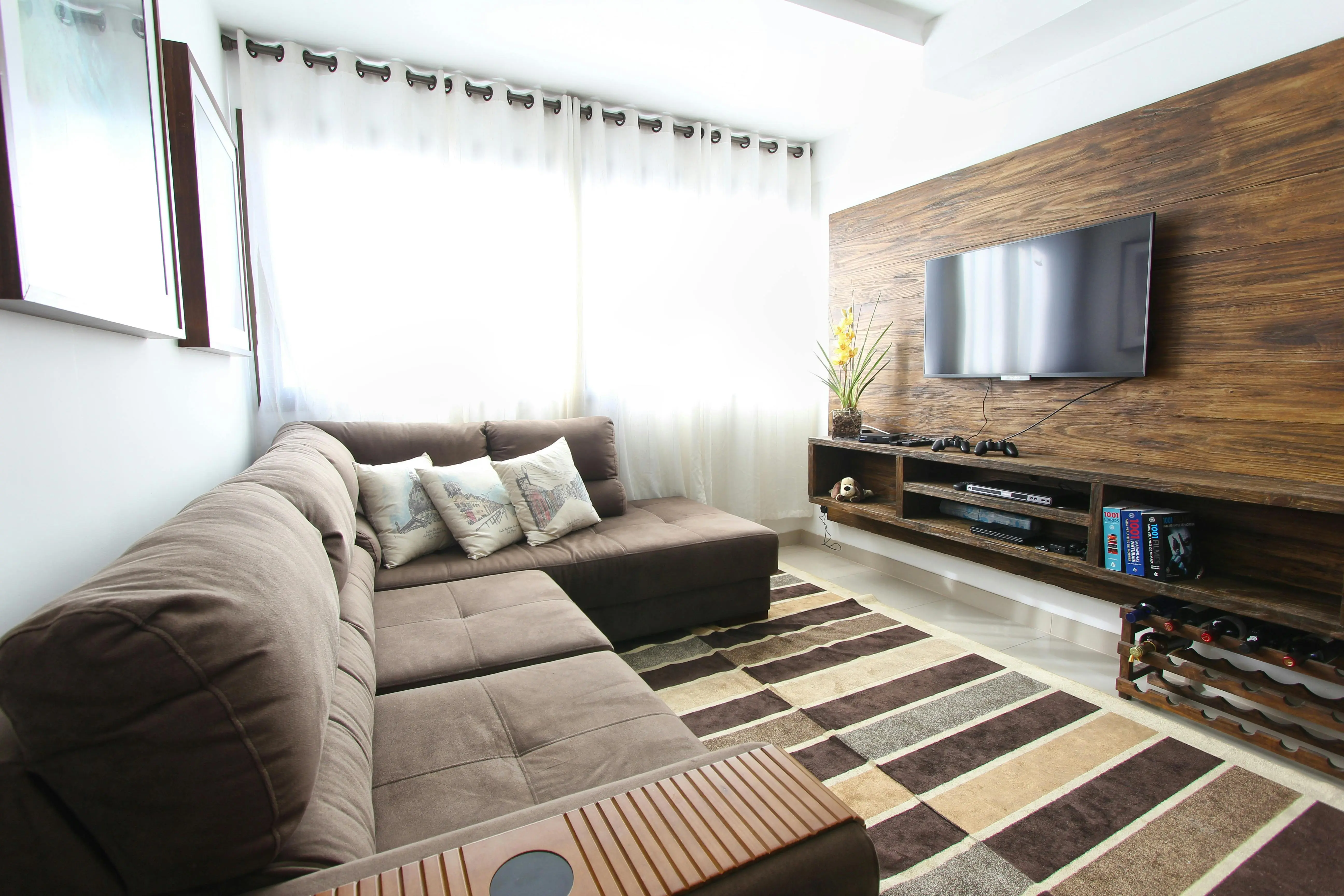 A brown sectional couch sits in front of a wooden-panelled wall. A TV is mounted on the wall, with a small shelf underneath filled with books and video game controllers.
