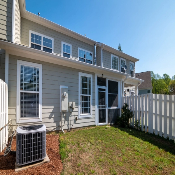 a sunlit backyard view of a modern grey house with white fencing and AC unit.