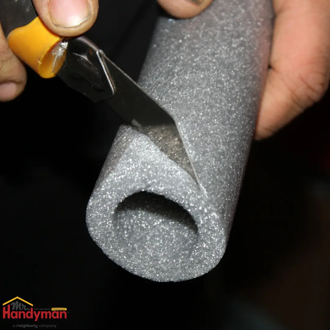 Close-up of hands using a utility knife with a yellow handle to cut a piece of gray, cylindrical foam pipe insulation.