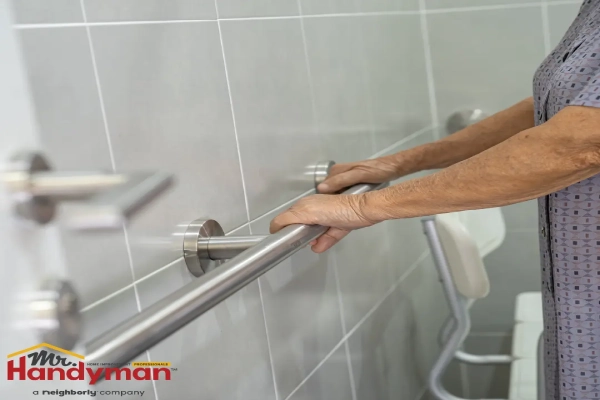 An elderly person using a stainless steel grab bar in a tiled bathroom to assist with mobility.