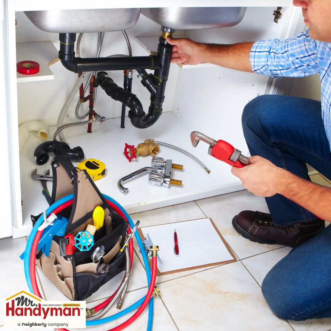 A plumber working under a kitchen sink, using a pipe wrench to adjust plumbing connections.