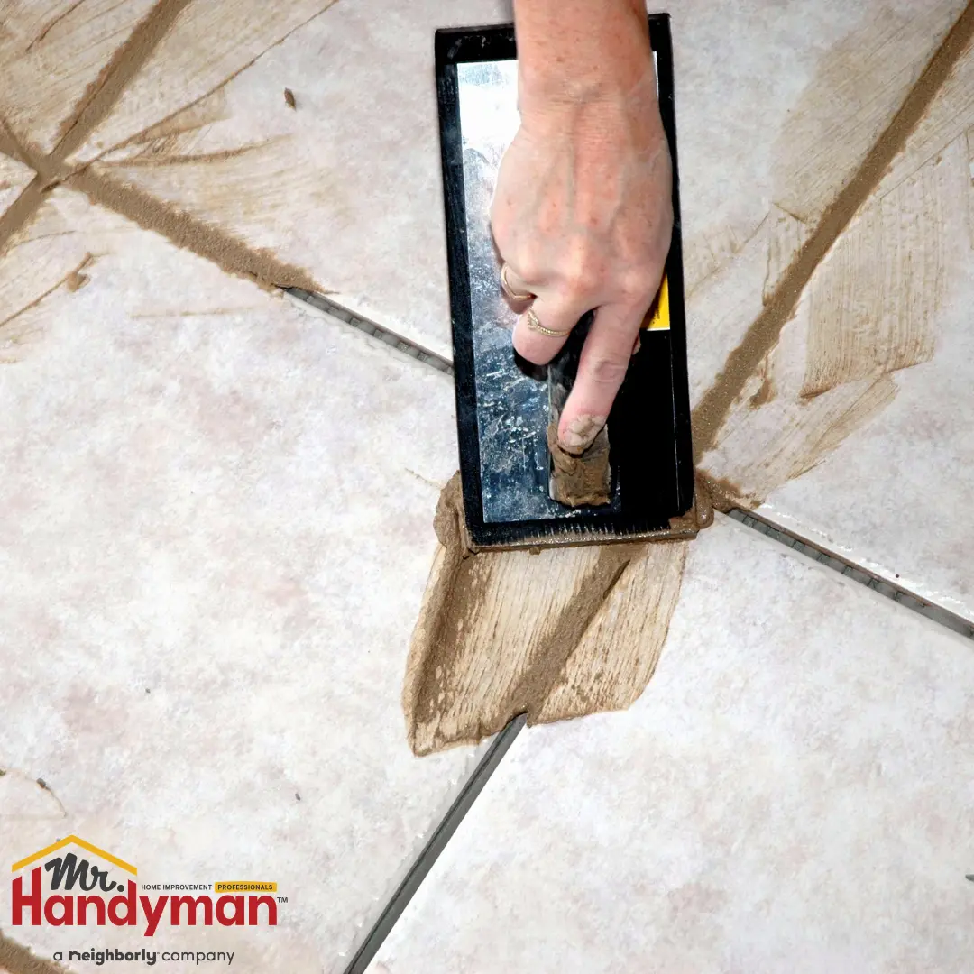 A technician using a trowel to apply and smooth tile grout along the joints of a tiled floor.