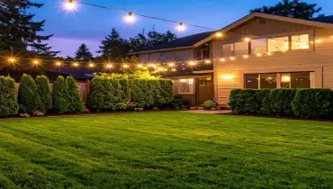 A cozy backyard at twilight with string lights, green grass, trimmed hedges, and a charming house.