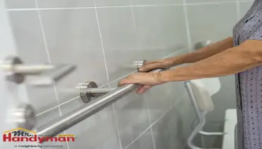 An older adult holds a stainless steel grab bar in a tiled bathroom.