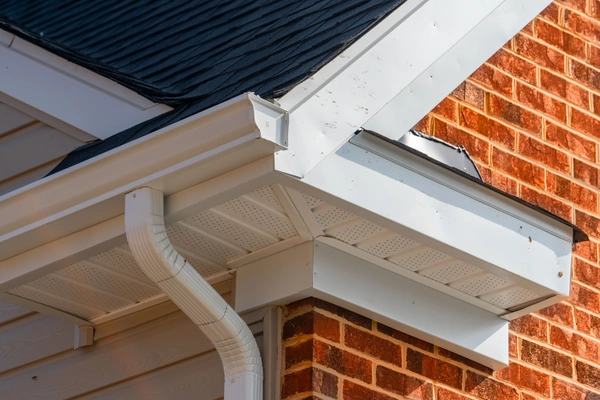 Close-up of a roof corner with a gutter and downspout against a brick wall.