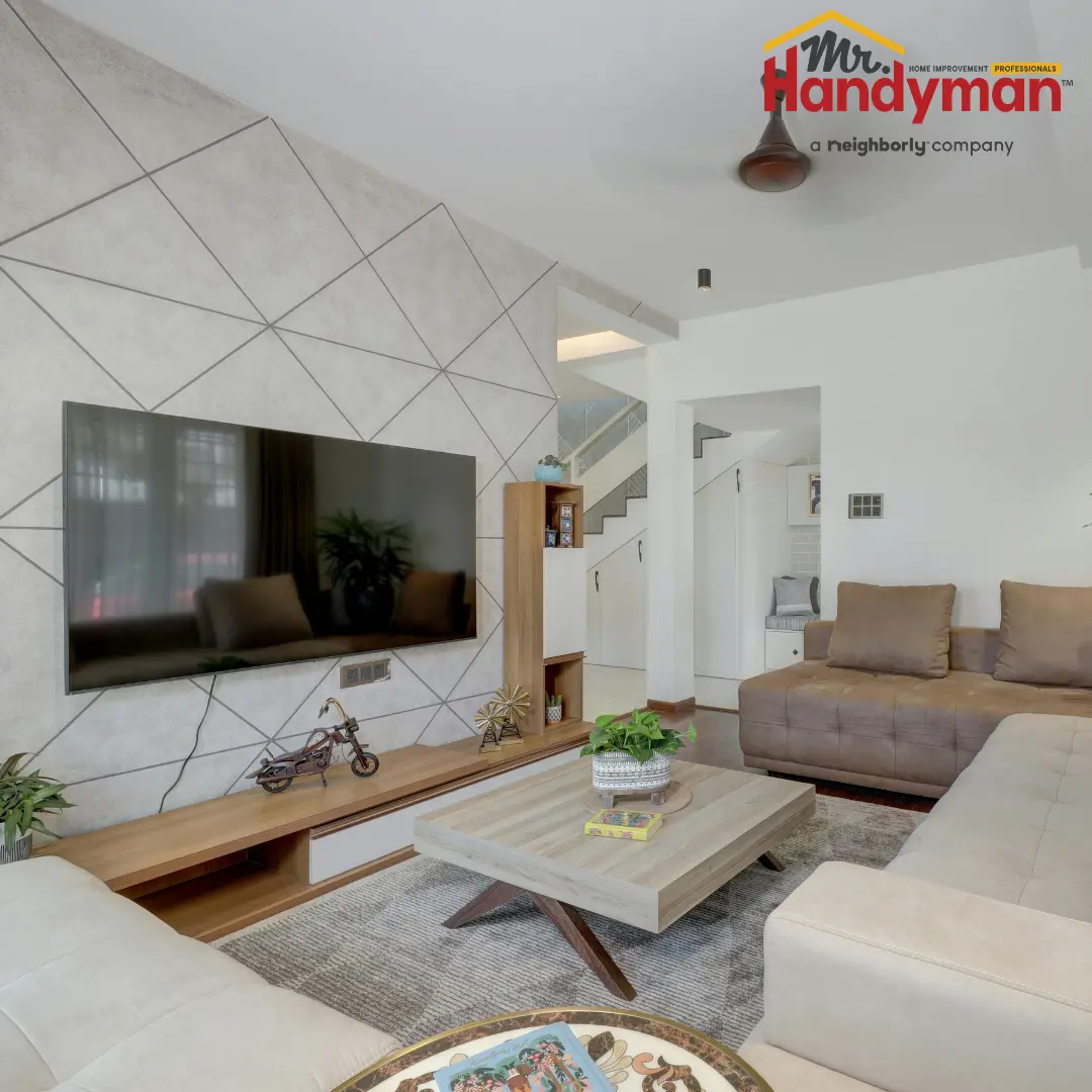A modern living room designed by Mr Handyman, featuring a geometric-patterned accent wall, a large wall-mounted television, and a mix of neutral and earth-toned furniture.