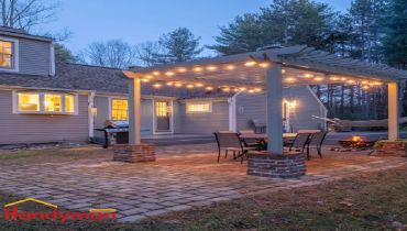 A cozy backyard patio area with string lights, a stone pathway, and a seating arrangement near a fire pit, surrounded by trees.