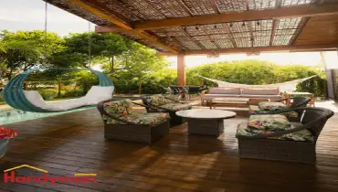 A cozy outdoor patio featuring a hanging chair, a sofa, and several chairs with floral patterns, surrounded by greenery.