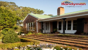 A charming brick house with a green roof, white-framed windows, and a covered front porch, surrounded by lush gardens and blooming flowers.