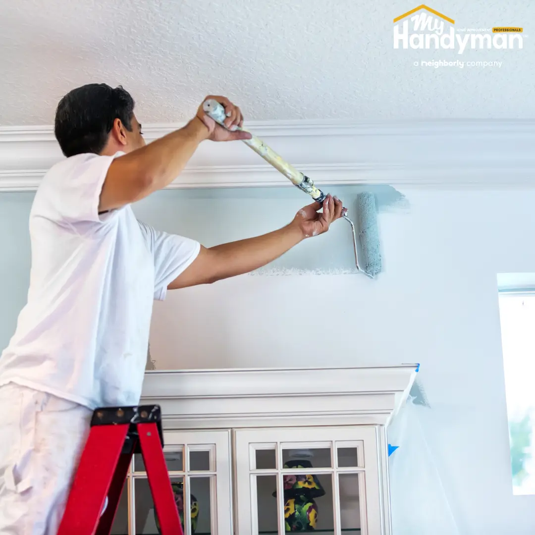 Person standing on a ladder, using a paint roller to paint a light-colored wall.