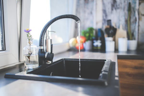 Photo of a kitchen sink and faucet that is running with water flowing out of it. There is a glass vase with a singular purple flower near the faucet.