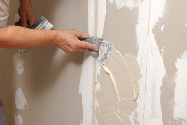 A person's hands using a putty knife to apply joint compound over mesh tape.