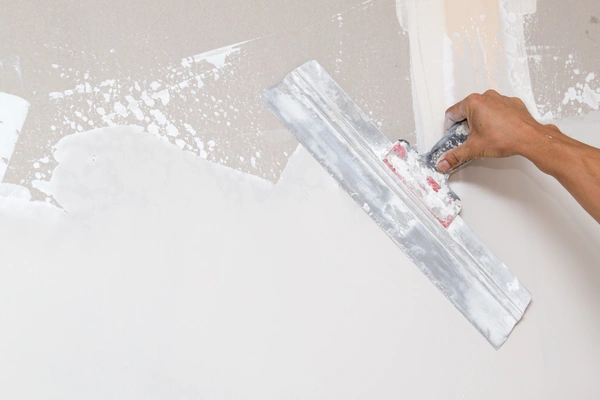 A hand using a wide drywall taping knife to smooth joint compound over a wall surface.