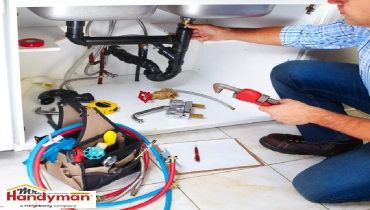 A plumber working under a kitchen sink, using a pipe wrench to adjust plumbing connections.