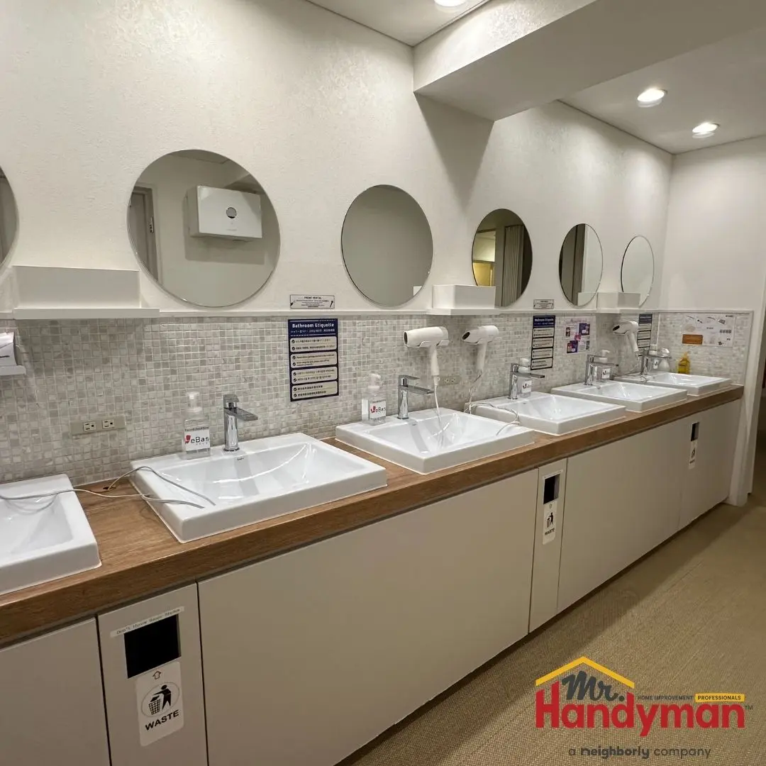 A row of modern public restroom sinks with square basins, mounted mirrors, and built-in hand dryers.