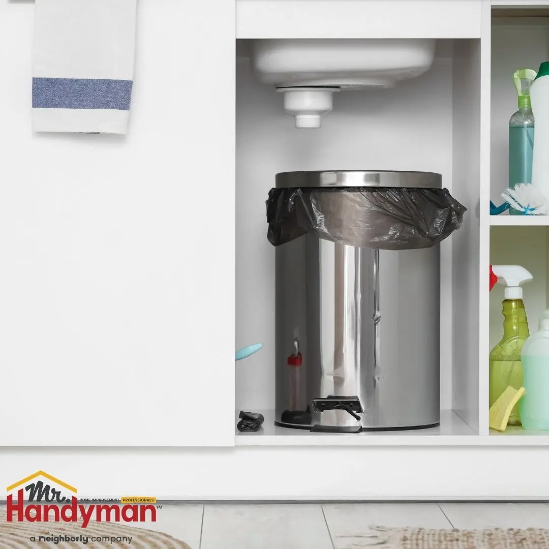 Mr. Handyman under-sink trash can installed in a white cabinet, with a black plastic liner and a foot pedal for easy access.
