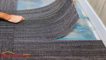 hands installing dark gray carpet tiles on a blue underlayment near a wall with white baseboard trim.