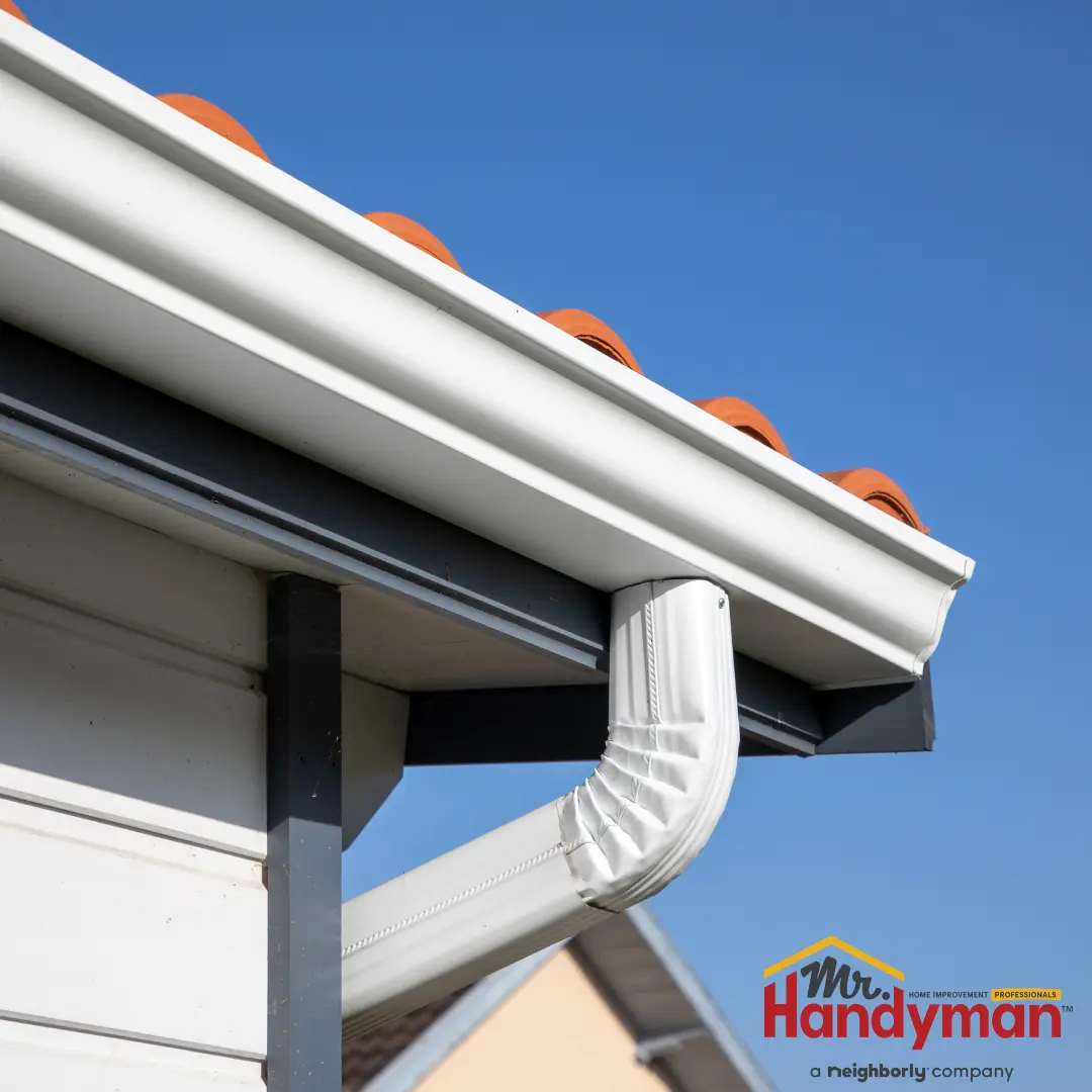 A close-up view of a white aluminum downspout and gutter system attached to the eaves of a house with a tiled roof.