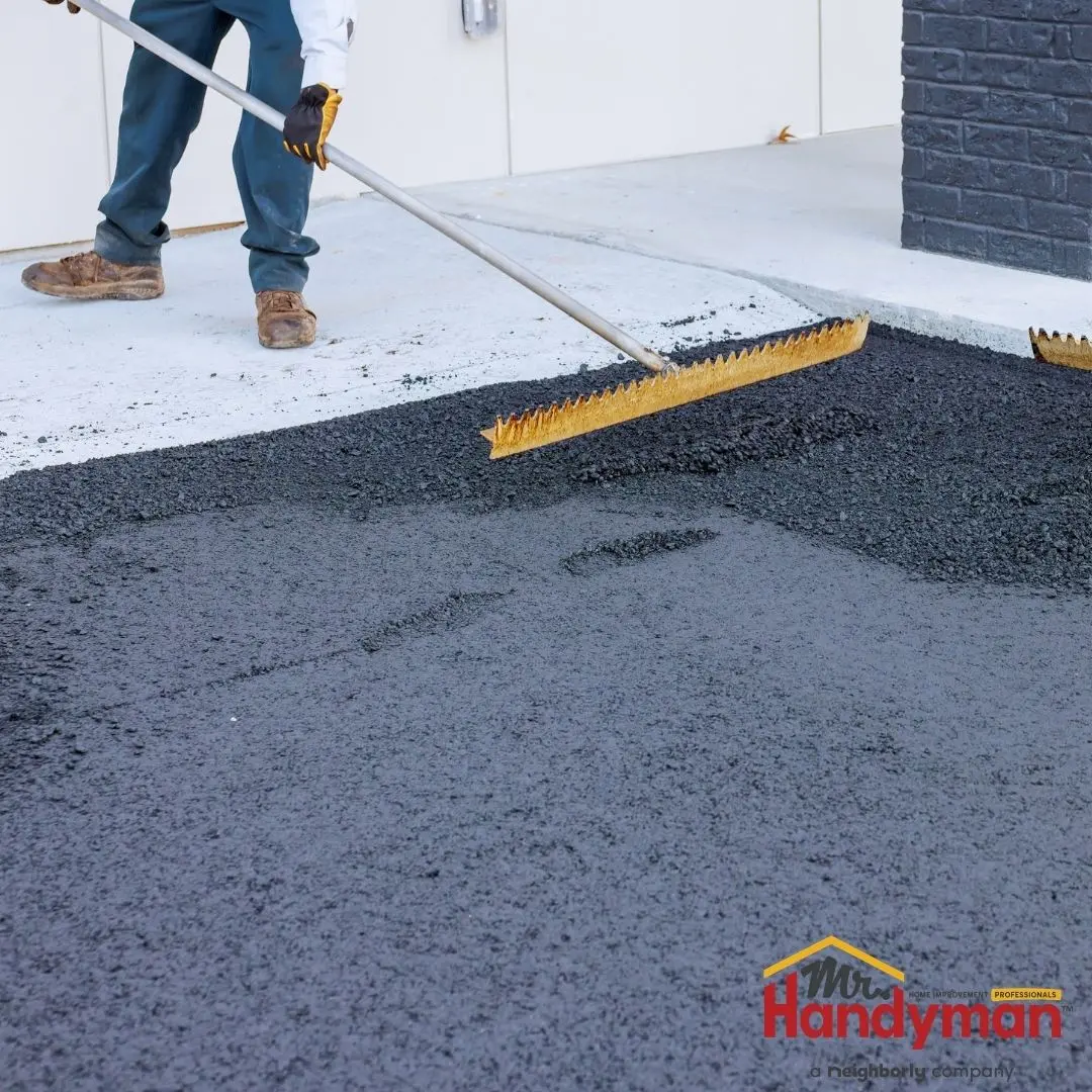A worker uses a yellow asphalt rake to smooth fresh black pavement on a driveway.