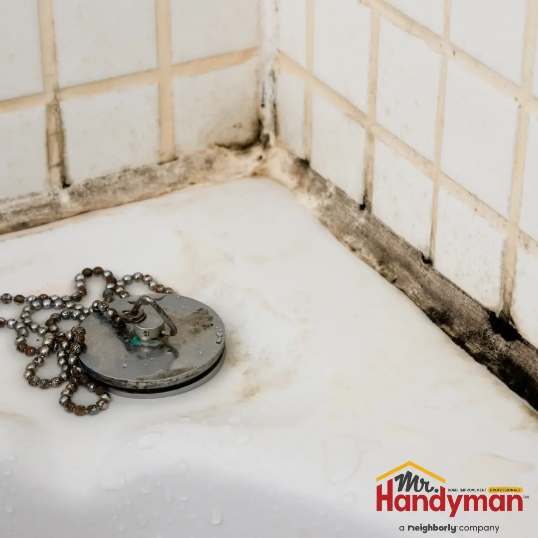 A bathroom shower drain with a chain drain cover, surrounded by white tiled walls and visible mold growth along the grout.