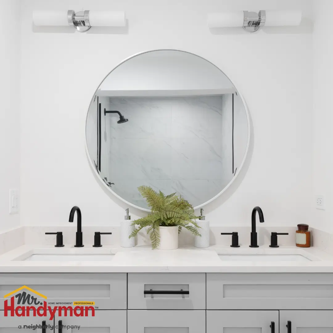 A modern bathroom vanity featuring a round mirror framed in white, flanked by two black faucets and a potted fern centerpiece.