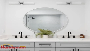 A modern bathroom vanity featuring a round mirror framed in white, flanked by two black faucets and a potted fern centerpiece.