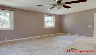 A room with beige walls, carpeted floor, two windows, and a ceiling fan with light fixture.