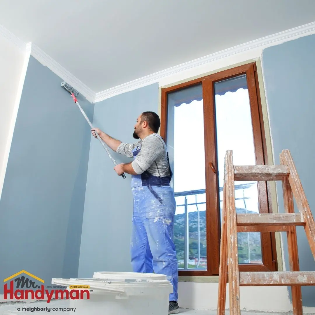 A man in blue overalls uses a long-handled roller to paint a wall light blue.