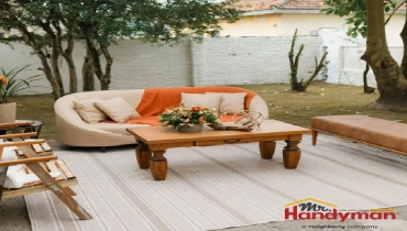 A cozy outdoor seating area featuring a curved cream sofa with an orange throw, a wooden coffee table adorned with flowers, and a striped rug.