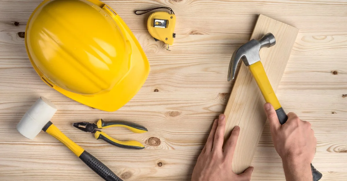 A handyman holding a hammer and a piece of wood near a collection of tools commonly used by a handyman in Lucas, TX, including a hard hat, mallet, pair of pliers and tape measure.