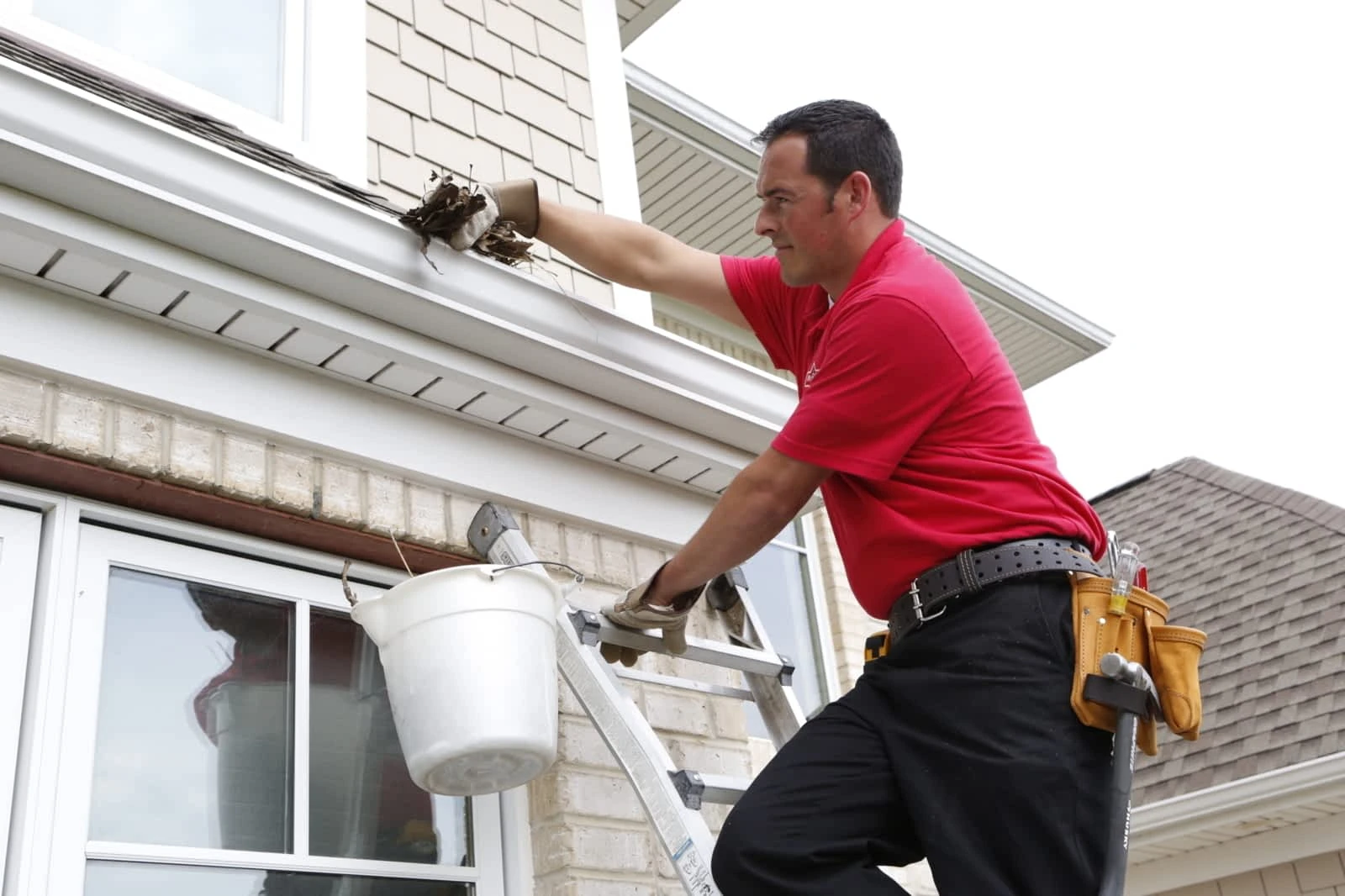 A Primrose handyman from Mr. Handyman standing on a ladder while cleaning leaves out of a gutter on the roofline of home.