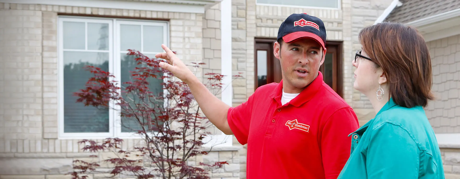 A handyman dressed in the Mr. Handyman uniform pointing to a home and speaking with a homeowner.