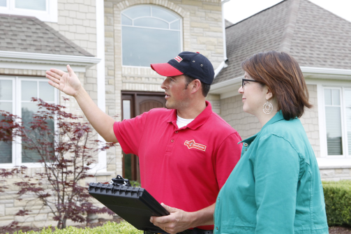 A handyman dressed in a Mr. Handyman uniform holding a clipboard and gesturing toward a home while speaking with a homeowner.