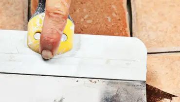 A handyman using a grout float to apply brown grout to the gaps between beige tiles on a floor.