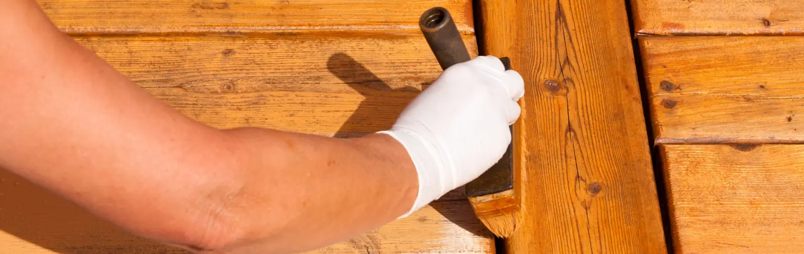 A person with a gloved hand applies stain to a deck with a brush.